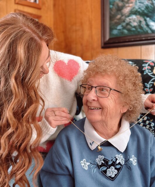 The author, Jami, who has red hair, is placing the gold cardinal necklace on her grandmother's neck while the smile at each other.