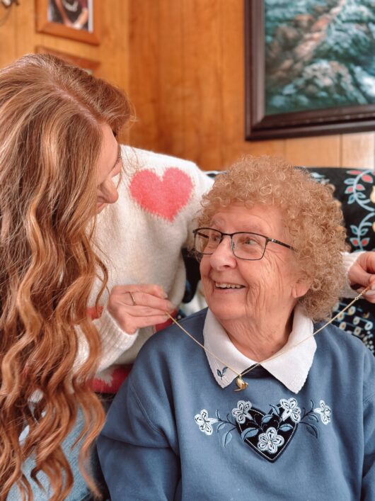 The author, Jami, who has red hair, is placing the gold cardinal necklace on her grandmother's neck while the smile at each other.