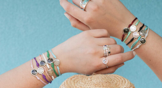 Two hands showcasing the Touch The World bracelets which include one sand and earth element each and colorful