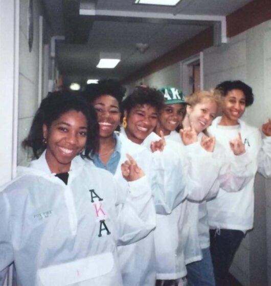 Six sorority sisters holding up their pinkies and smiling at the camera while wearing white coats.