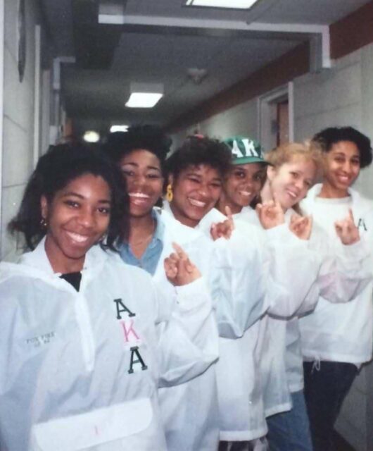 Six sorority sisters holding up their pinkies and smiling at the camera while wearing white coats.