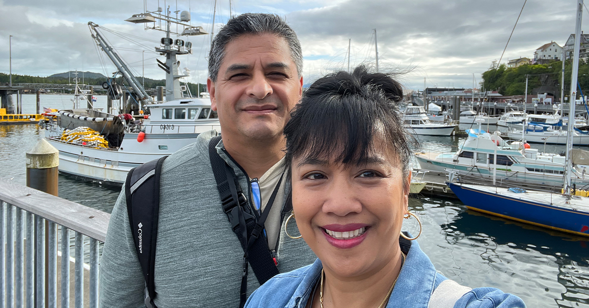 Couple stands with boats on their romantic trip to Alaska. Capture any travel memory in Dune Jewelry's Traveler Bracelet, a silver cuban chain link with customizable sand and earth elements.