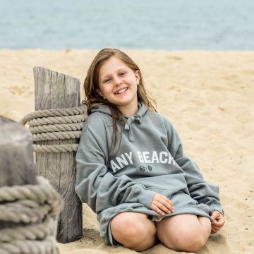 Girl smiling on the beach wearing a grey beach hoodie with the words any beach in white puff capital lettering.